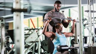 a photo of a woman working out on the lat pull down machine with a male trainer helping out