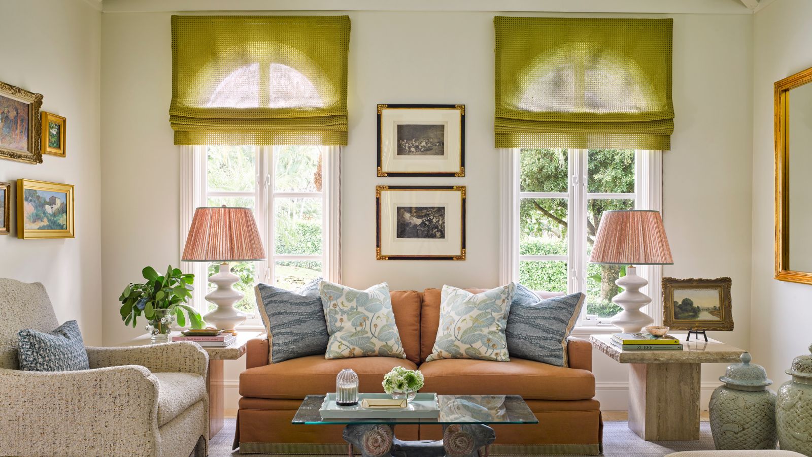 A large white living room with two tall arched windows with lime green sheer roman blinds, a terracotta-colored sofa with pale blue cushions, and two matching pink lampshades either side of the sofa. 