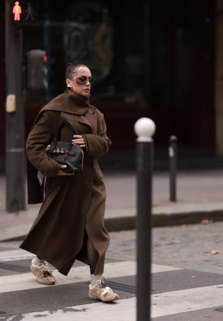 Anna Wein is seen wearing rimless oversized sunglasses with brown lenses, slim gold metal temples, and a gold metal nose bridge from Miu Miu; chunky squared earrings in gold with a black stone; an ankle-length brown wool trench coat featuring a double-button placket and notch lapels from Sandro; paired with a color-matching wide long scarf, also from Sandro; a rectangular black leather bag with gold details and several bag charms from Miu Miu; beige socks; and beige suede sneakers with fluffy lining and two different shoelaces in cream white cotton and brown leather from Miu Miu x New Balance during Paris Fashion Week on September 29, 2024 in Paris, France.