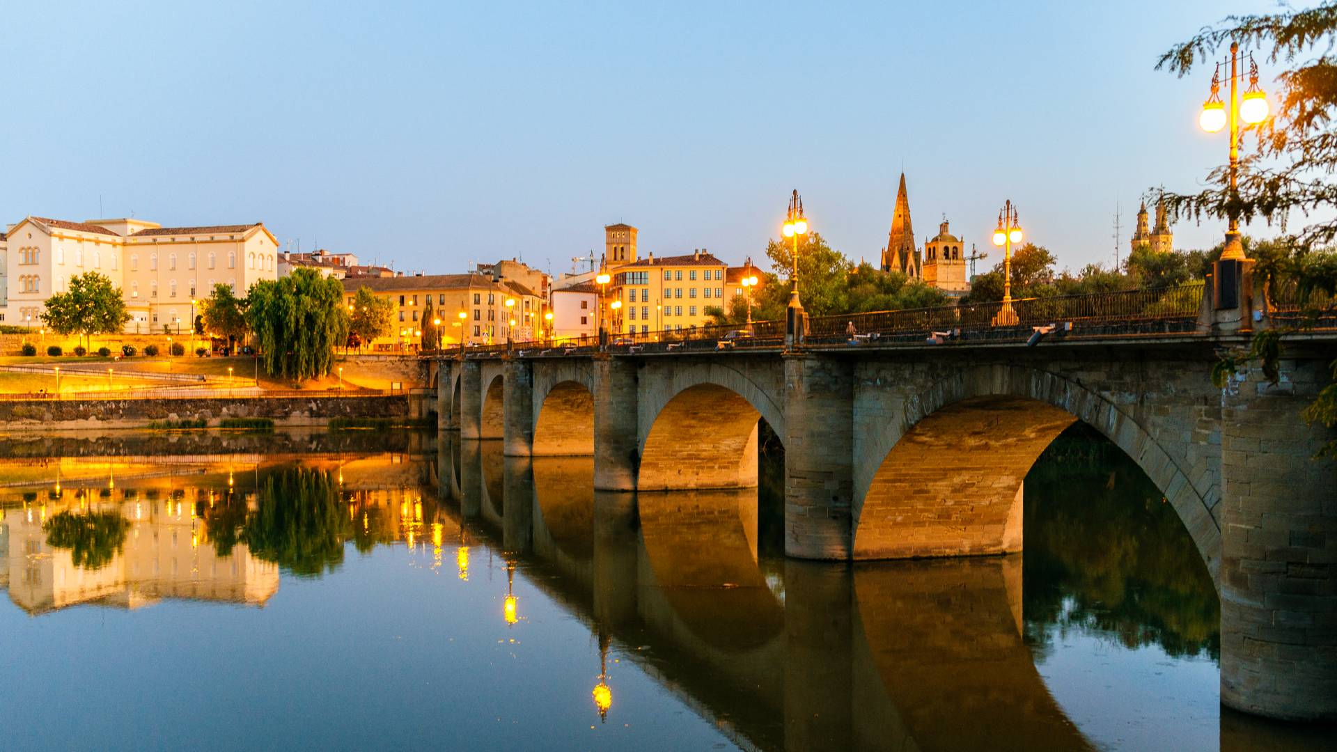 A stone bridge with arches spans a calm river at dusk, reflecting city buildings, trees, and church spires illuminated by warm streetlights against a clear evening sky.
