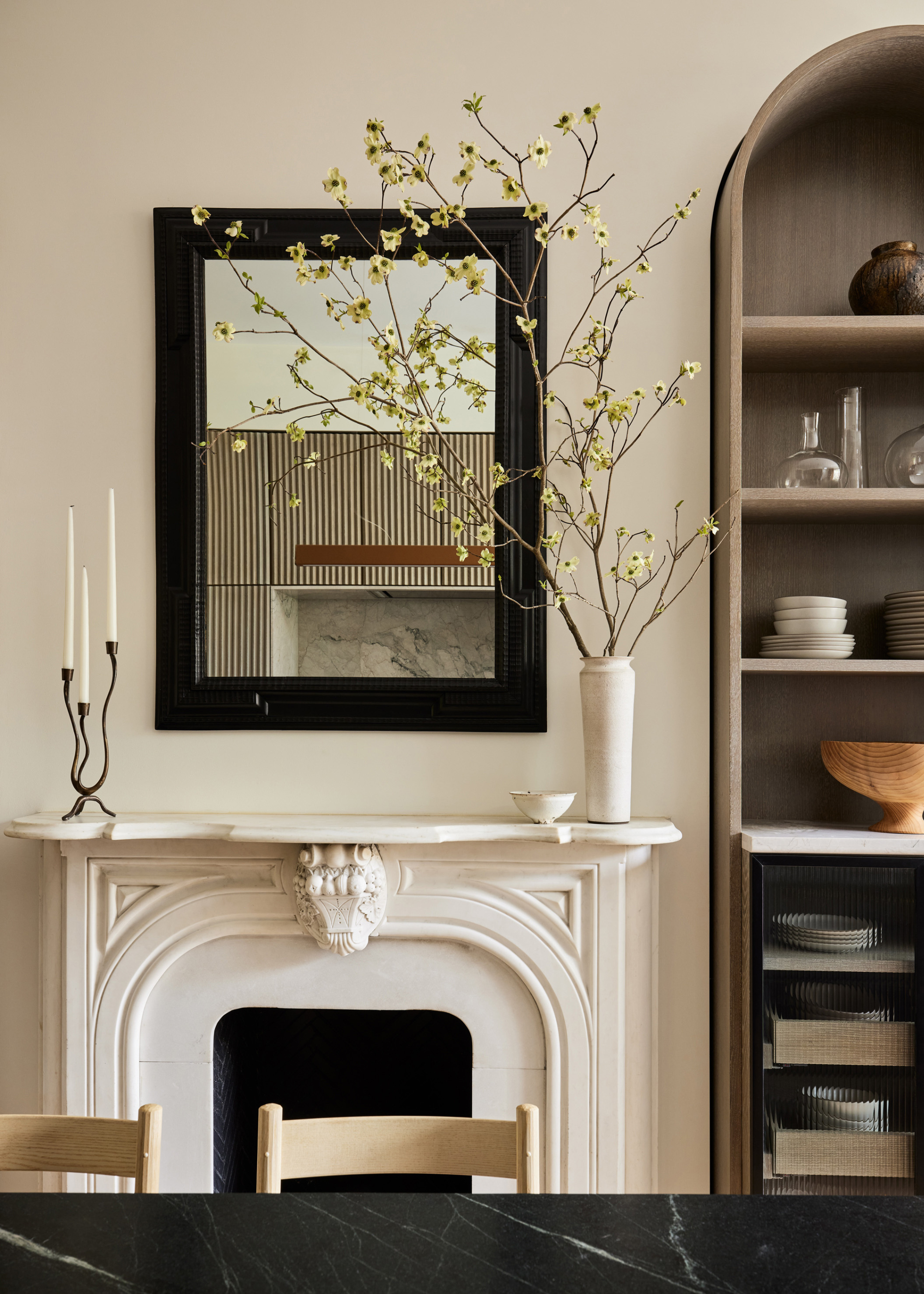 A dining room mantel with a candleholder, a vase of stems, a framed mirror, an arched display storage case, and a stone table