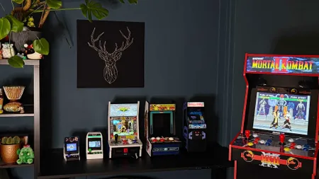 Line up of arcade machines in living room with green wall including Neo Geo Mini, Sega Astro City Mini, Evercade Alpha, Arcade Classics Centipede, Bubble Bobble Quarter Arcades, and Arcade1Up Mortal Kombat 2 machine (from left to right).