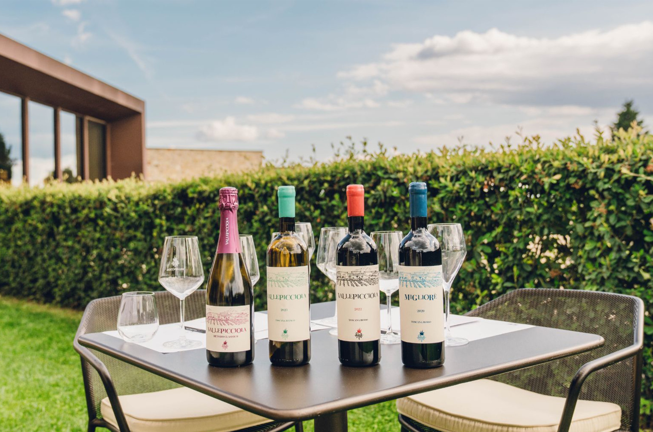 Four bottles of wine: sparkling, white and two red, on a table outside with wine glasses