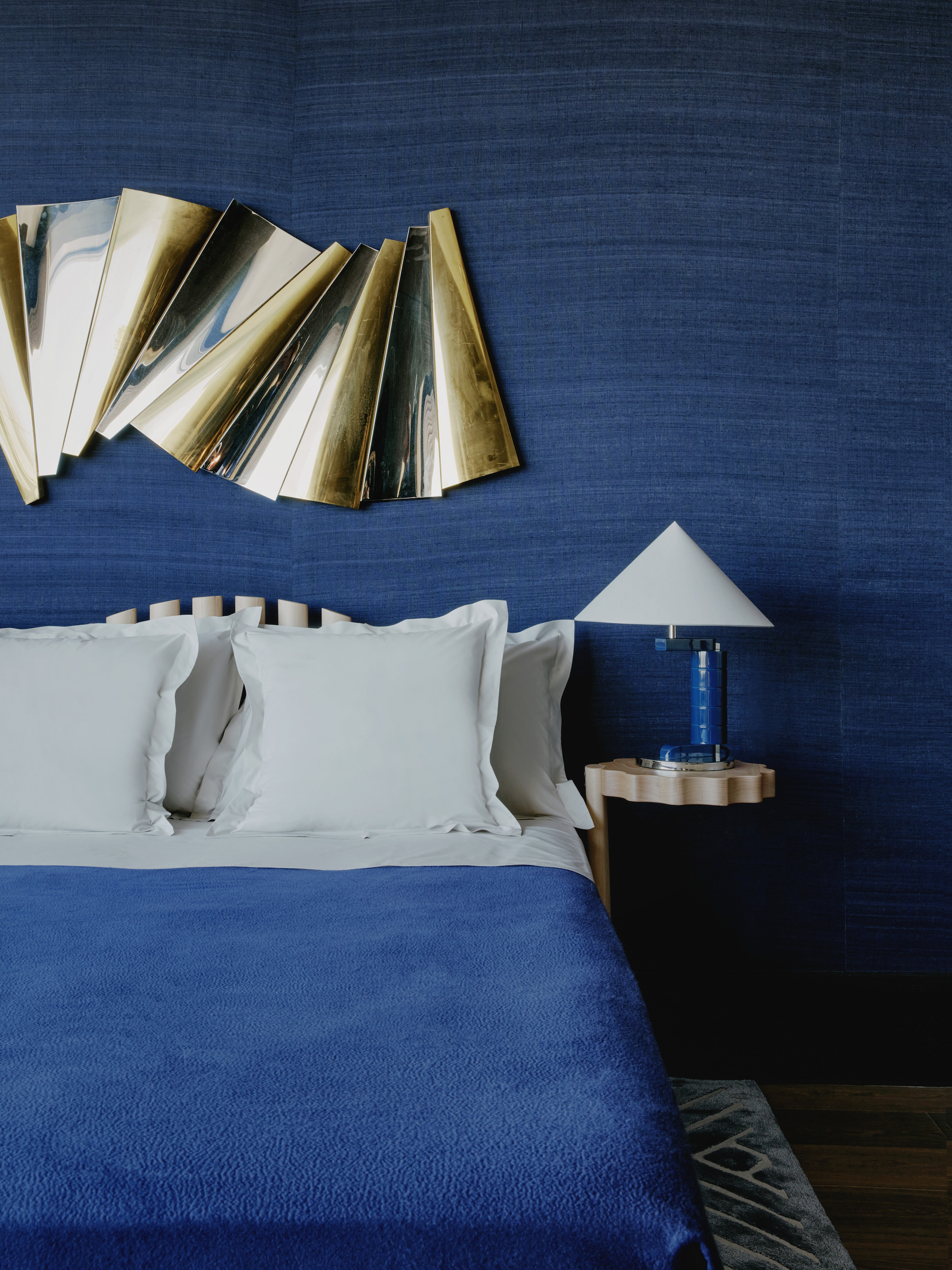 A deep, sea blue bedroom with matching blanket and bedside lamp. A gold, art piece hangs above the headboard.