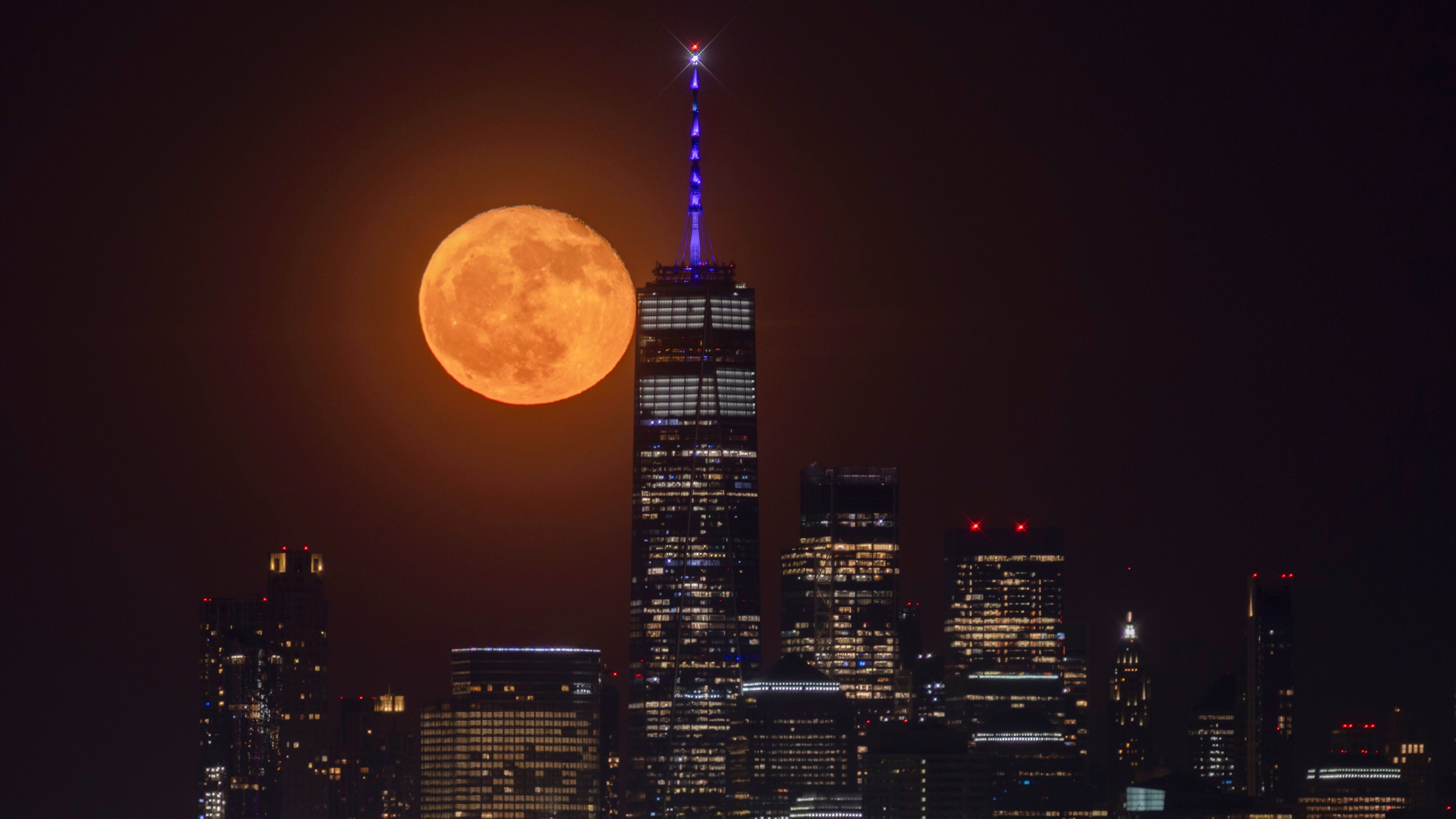 An orange full moon glows to the left of a skyscraper looming above a city skyline at night