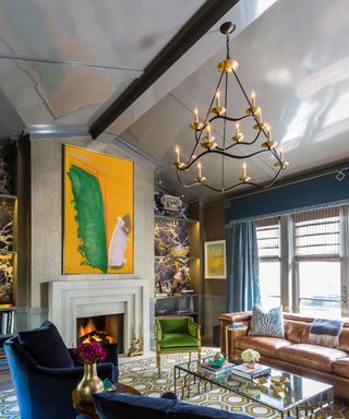Grand living room with gray gloss painted vaulted ceiling, black metal chandelier, geometric patterned rug, two blue velvet armchairs, green velvet chair, brown leather sofa, glass coffee table, blue curtains with pelmet, large yellow artwork above fireplace