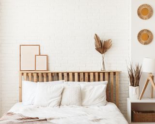 Tidy white bedroom with wooden headboard with dry gold palm leaves in a glass vase and two photo frames on it. Stylish trendy decoration with copy space.