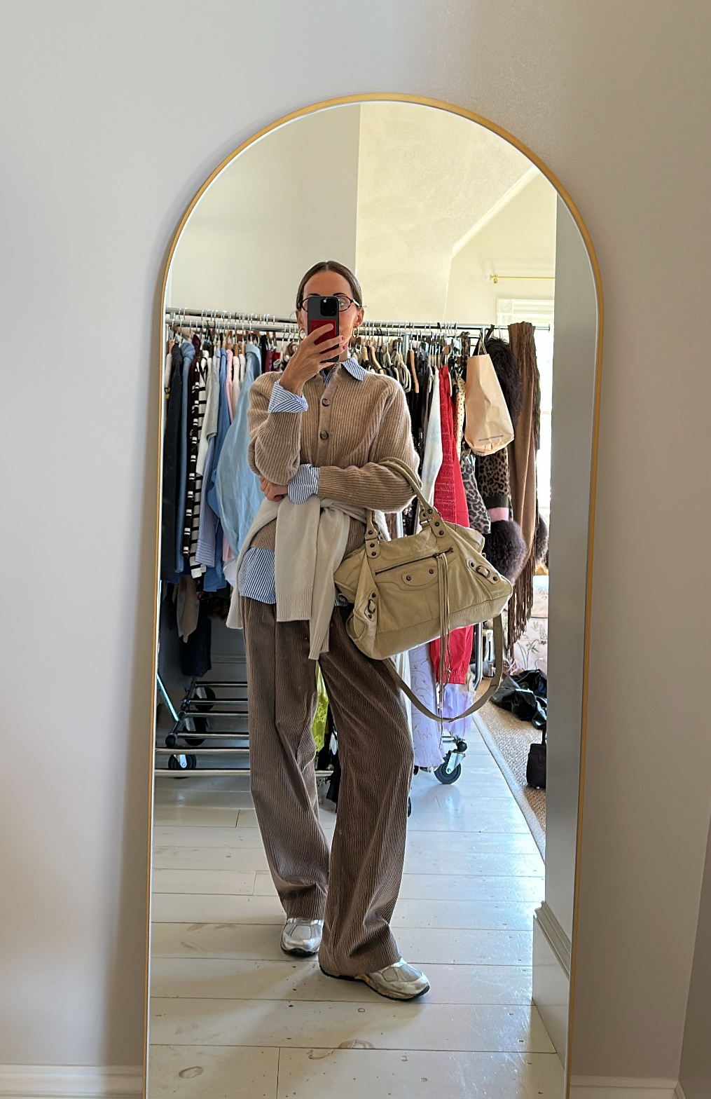 Larissa Mills wearing a blue striped shirt, tan cardigan sweater, white sweater around her waist, brown pants, white sneakers, and a tan Balenciaga city bag