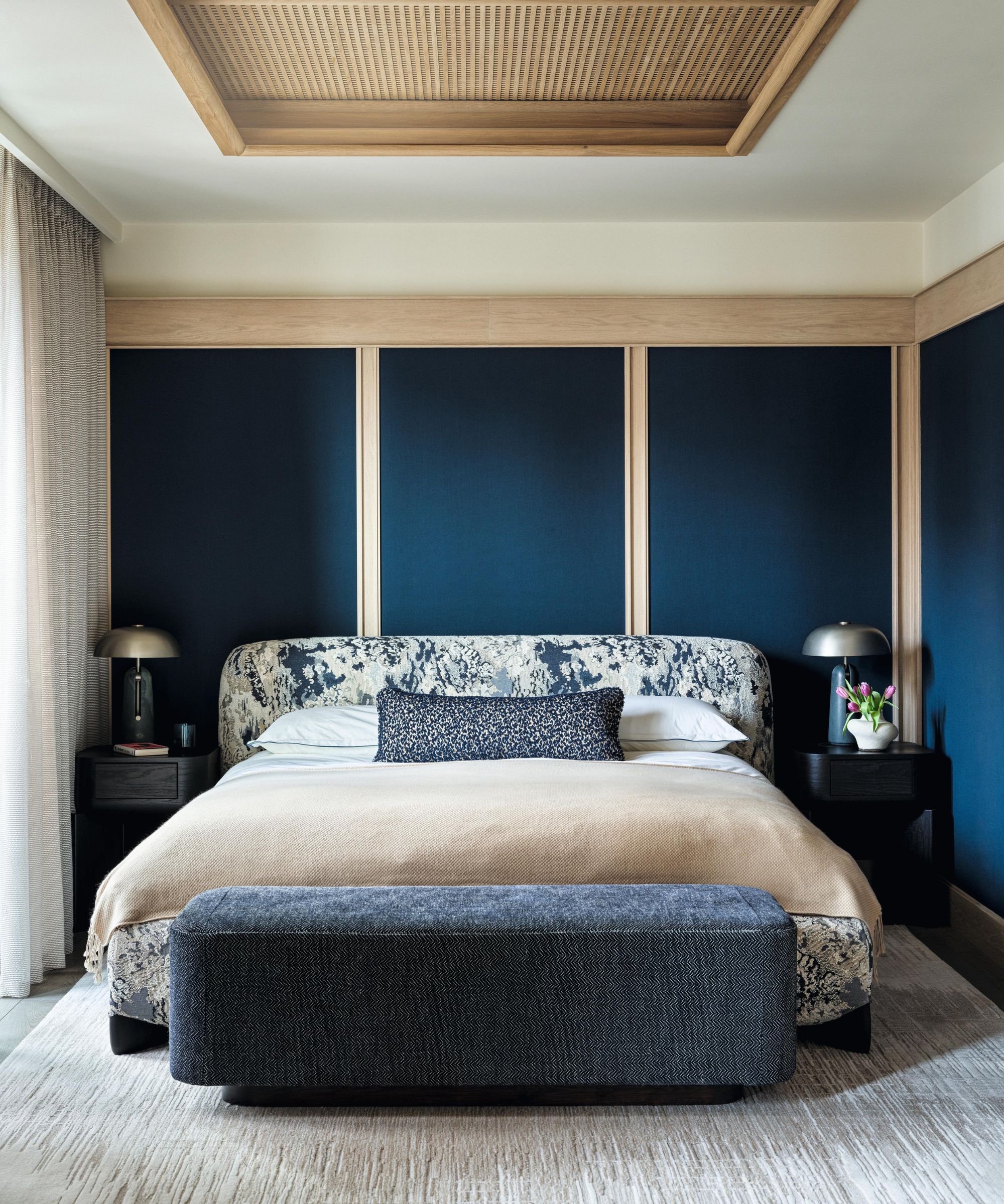 a paneled main bedroom with navy and wooden panels, a ceiling wooden area, floral upholstered bed, with a blue ottoman at the base of the bed
