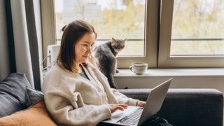 A woman sat on the sofa next to a cat working on her laptop