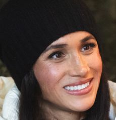 Meghan Markle smiling and wearing a black winter hat and white puffer coat