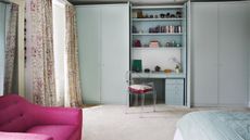 Small bright bedroom with hidden vanity in the built-in closet. There is shelving above the vanity and clear plastic chair. The room has a large window with floor length floral curtains and a bright pink accent chair