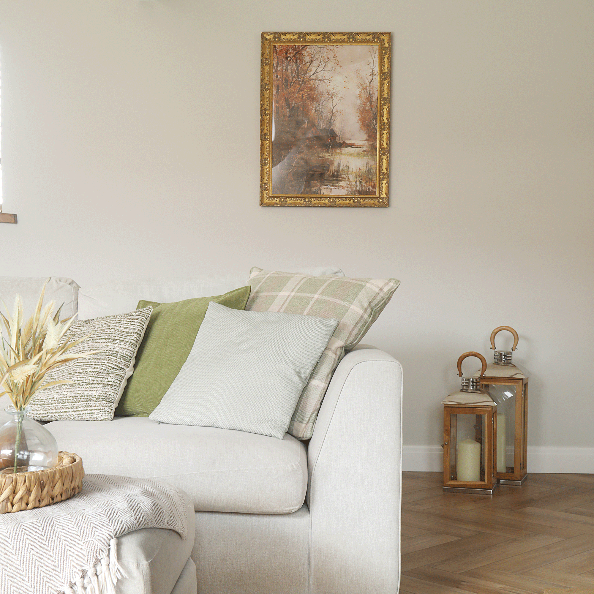 a living room with a cream sofa and matching footstool with a couple if large lanterns with pillar candles and a gold framed artwork on the wall
