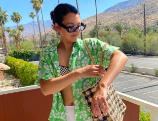 Woman on balcony in Palm Springs wearing a floral shirt, bikini, and white skirt with palm trees in the background.