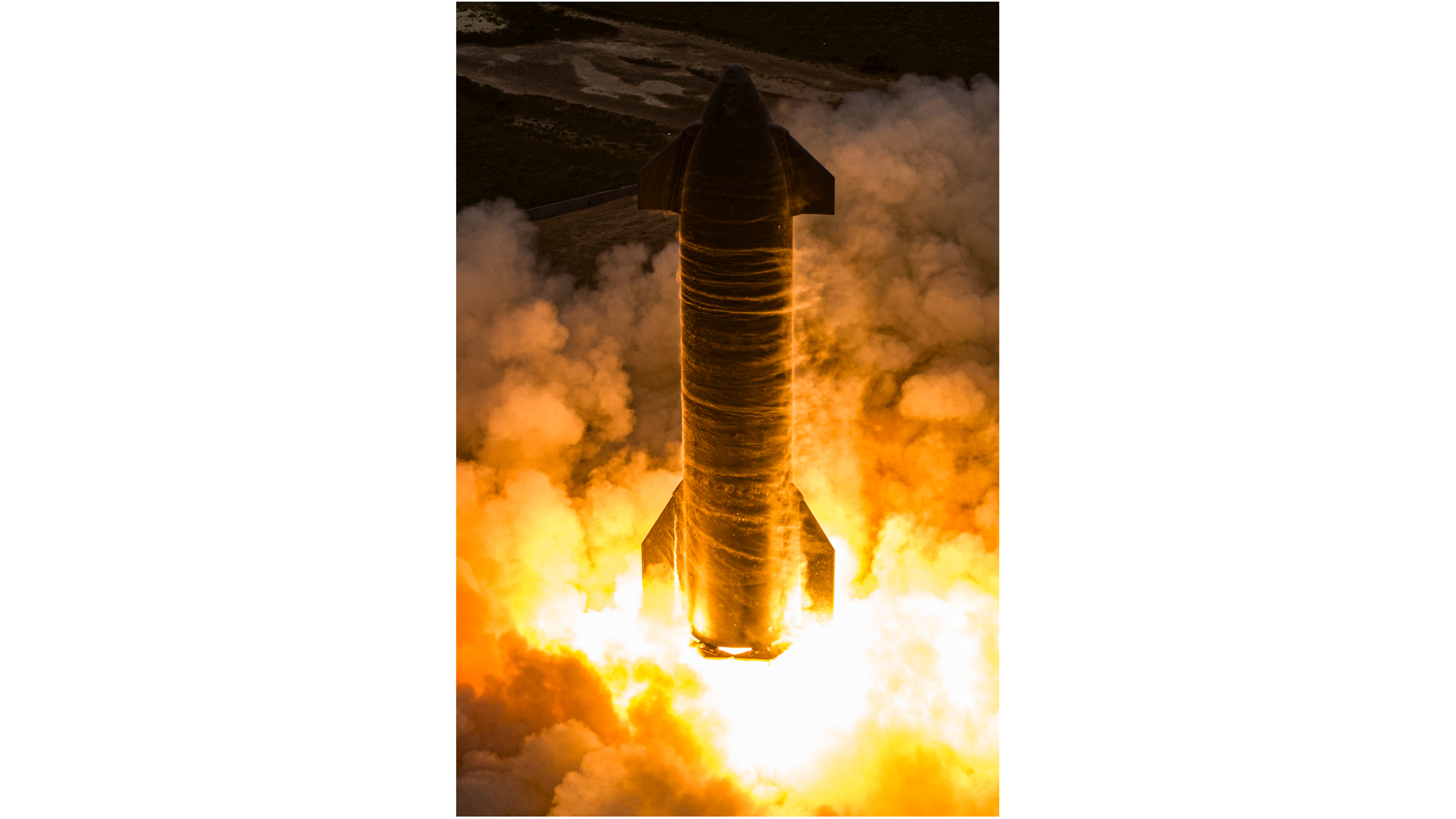 Foto lateral, que muestra un cohete cilíndrico plateado con llamas anaranjadas que salen de su base.