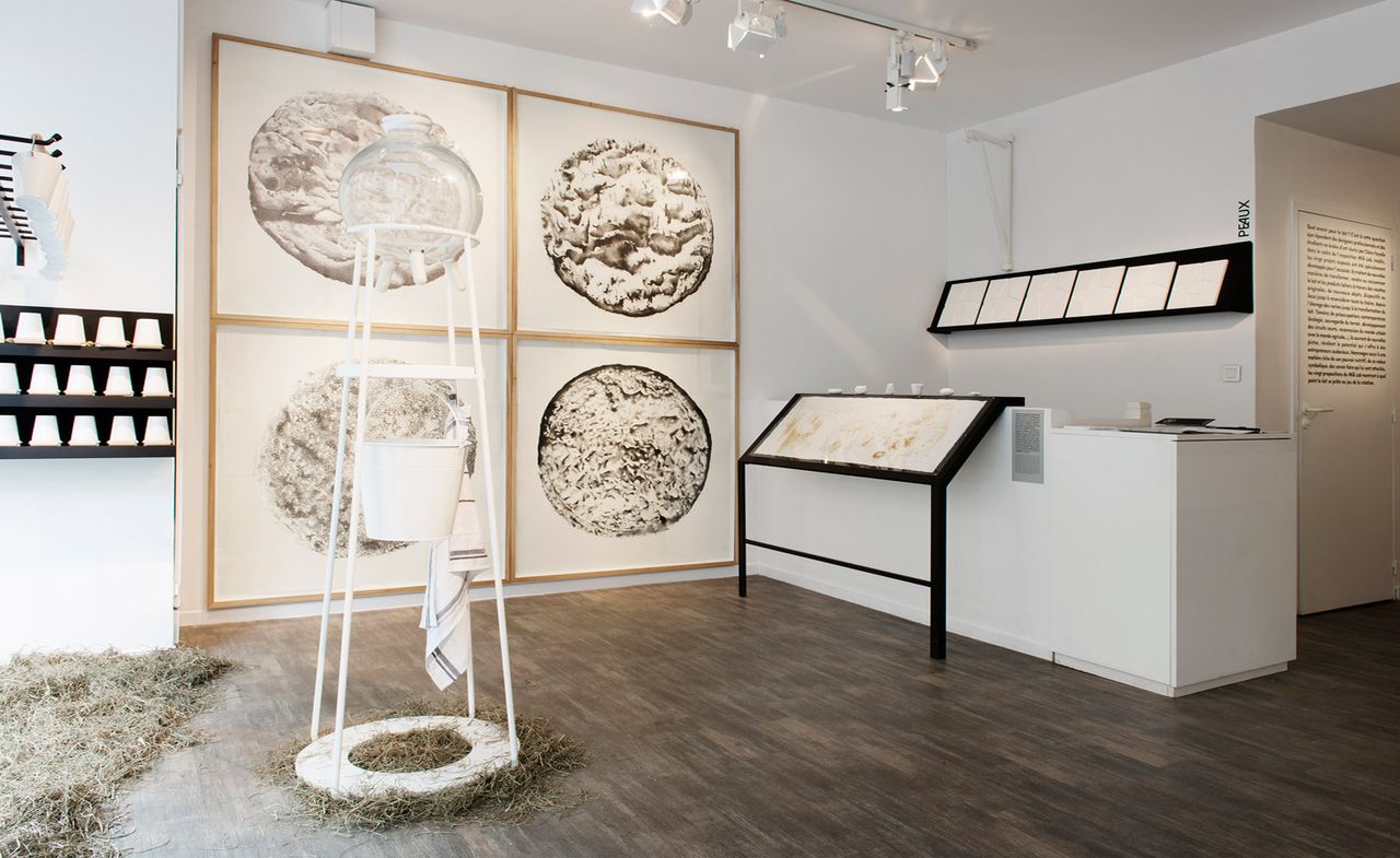 An art display room featuring white walls and ceiling with dark wood flooring. In the middle is a white cone shaped structure with synthetic udders suspended above a white traditional milk pail &amp; white towel which is hanging off structure. On the wall is a quad display of 4 curdles cheeses.