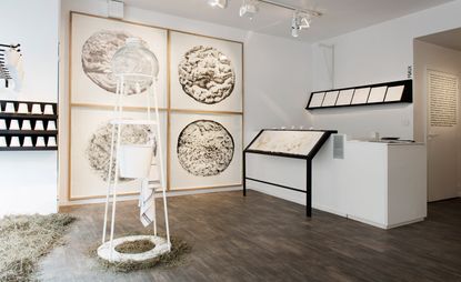 An art display room featuring white walls and ceiling with dark wood flooring. In the middle is a white cone shaped structure with synthetic udders suspended above a white traditional milk pail & white towel which is hanging off structure. On the wall is a quad display of 4 curdles cheeses.