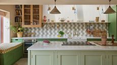 eclectic large green kitchen with a bench seat and checkerboard fabric, lighter green kitchen island with two pendant lights above, striped tiled backsplash and glass fronted wall cabinets