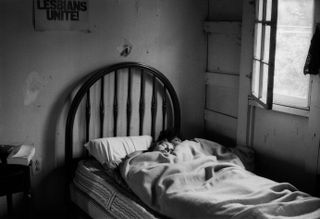 Black and white image of two women asleep in a bed. A picture on the wall has the words "LESBIANS UNITE!"