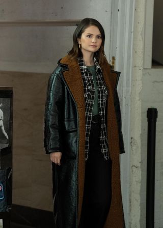 Selena Gomez wearing a leather trench coat lined with shearling, a plaid jacket, a green top, and black pants