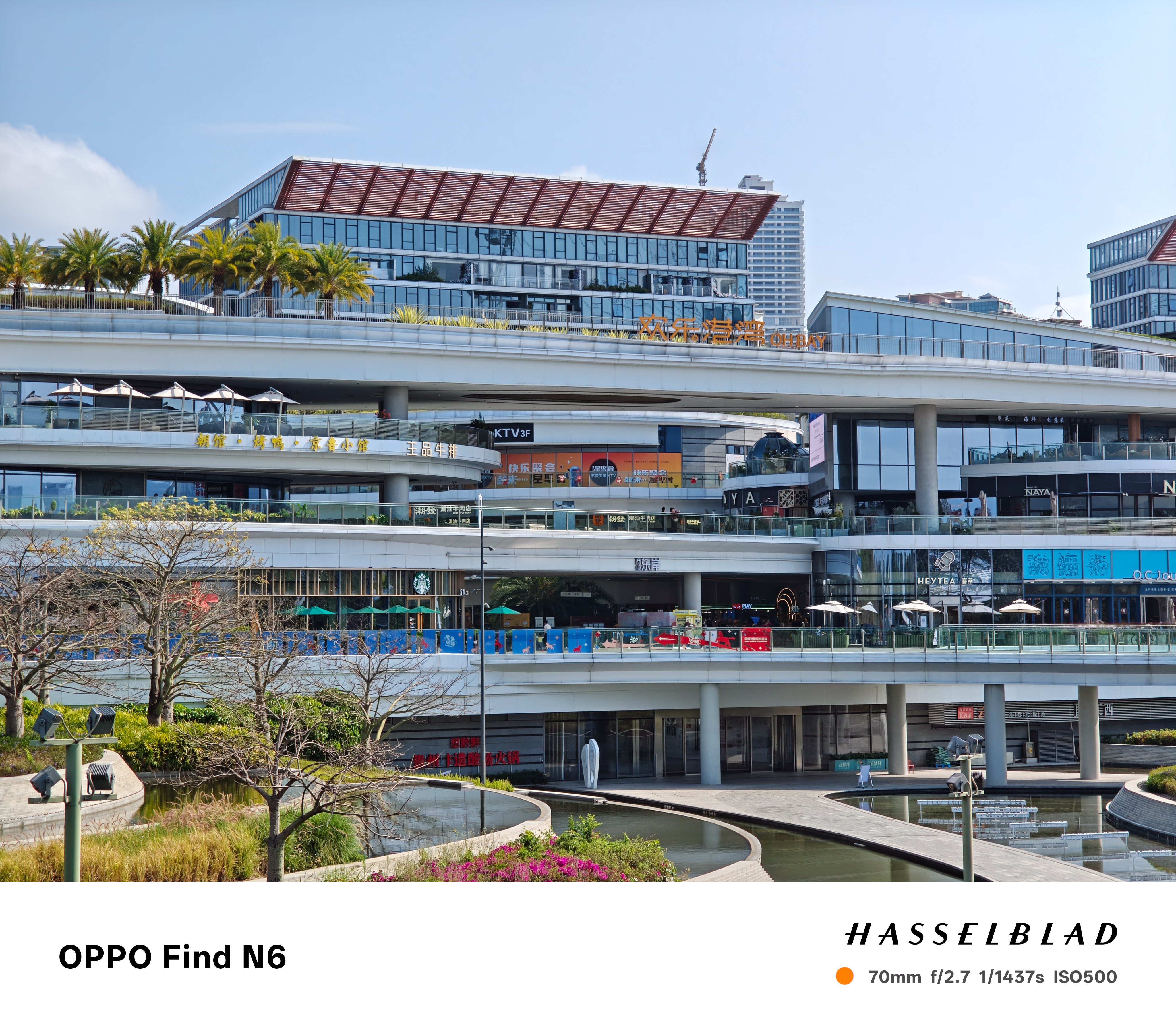 A telephoto shot of the upper levels of a white modern building, showcasing palm trees on a terrace and various storefronts. The perspective highlights the clean lines of the architecture and the transparency of the glass balconies.