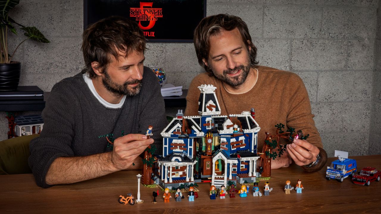 The Duffer Brothers hold up minifigures while sitting behind the Lego Creel House