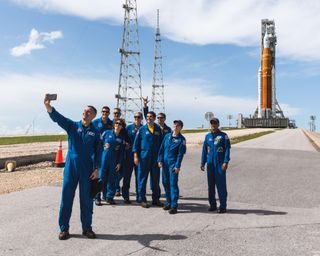 nine people in blue flight suits pose for a picture with an orange rocket looming over them in the background