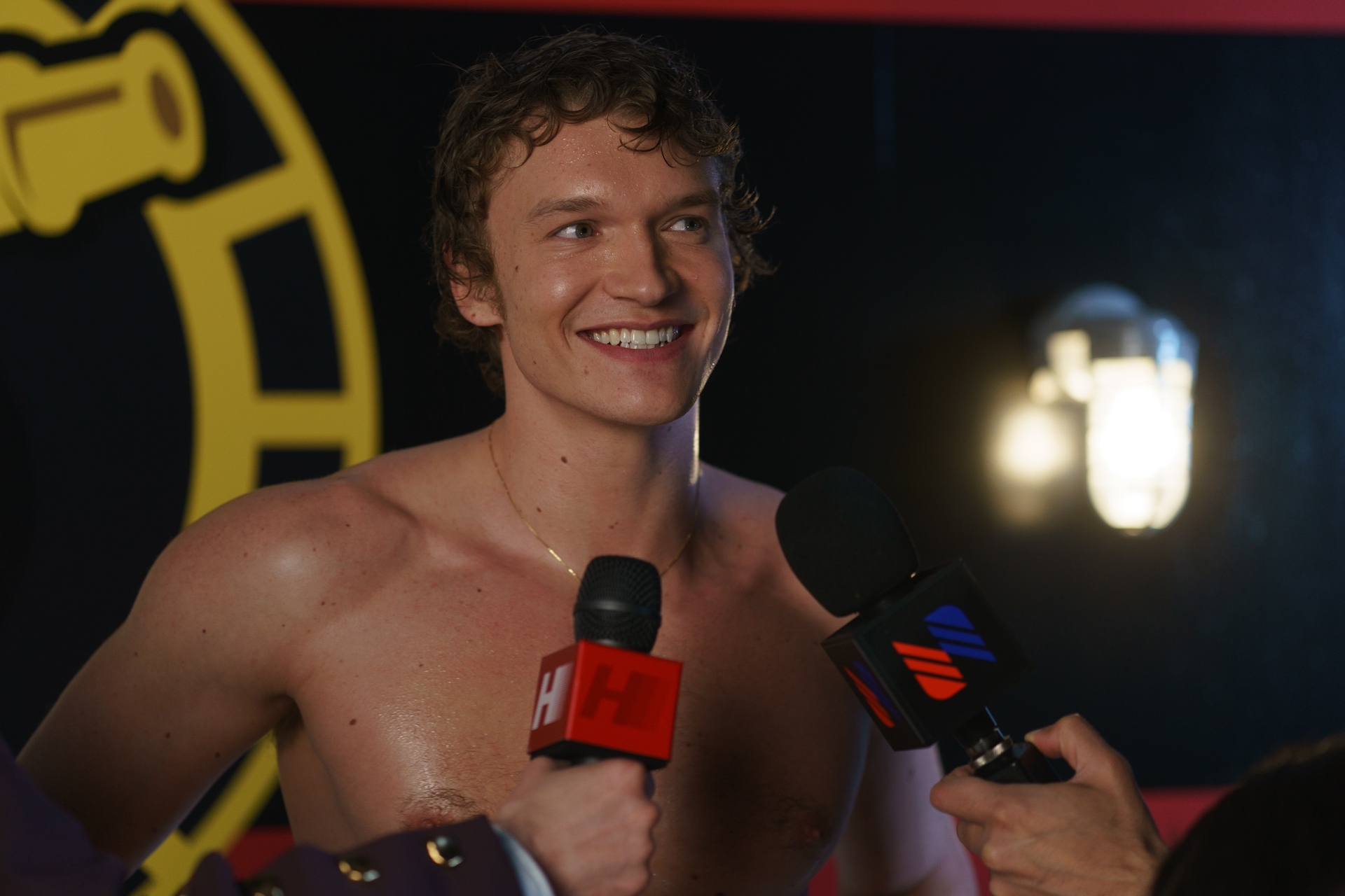 Connor Storrie as ilya shirtless and smiling as he answers reporter interviews and speaks into microphones in Heated Rivalry