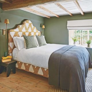 bedroom in Cotswold cottage with large upholstered bed, green painted walls and beams on ceiling, with bare floorboards