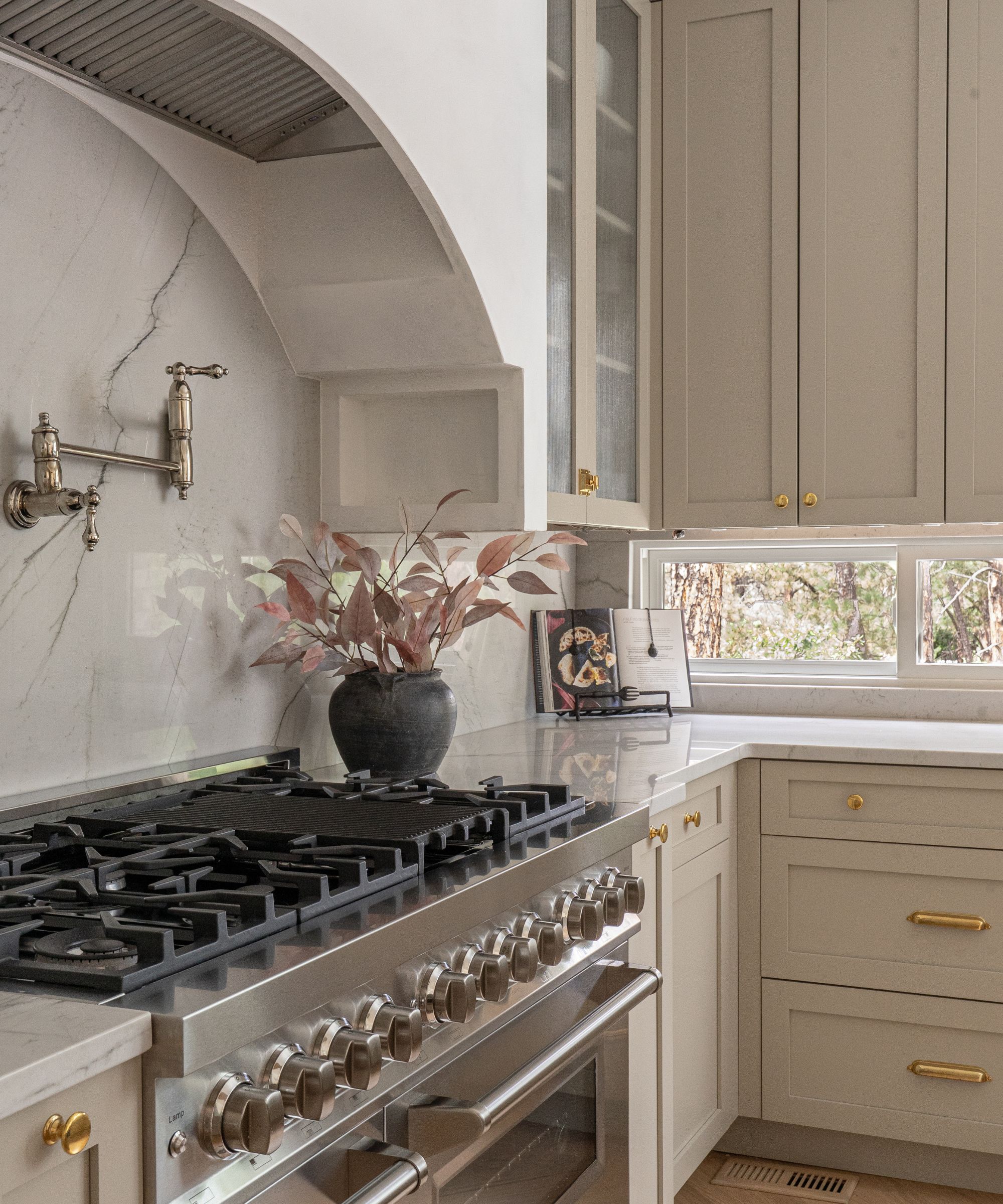 Kitchen with curved range hood painted white, Pashmina by Benjamin Moore painted cabinetry, gold hardware, a silver range oven, and a silver pot filler, with a vase of pink foliage.