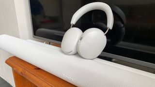 A white pair of Sonos Ace headphones on top of a white Sonos Arc Ultra soundbar in front of a TV.
