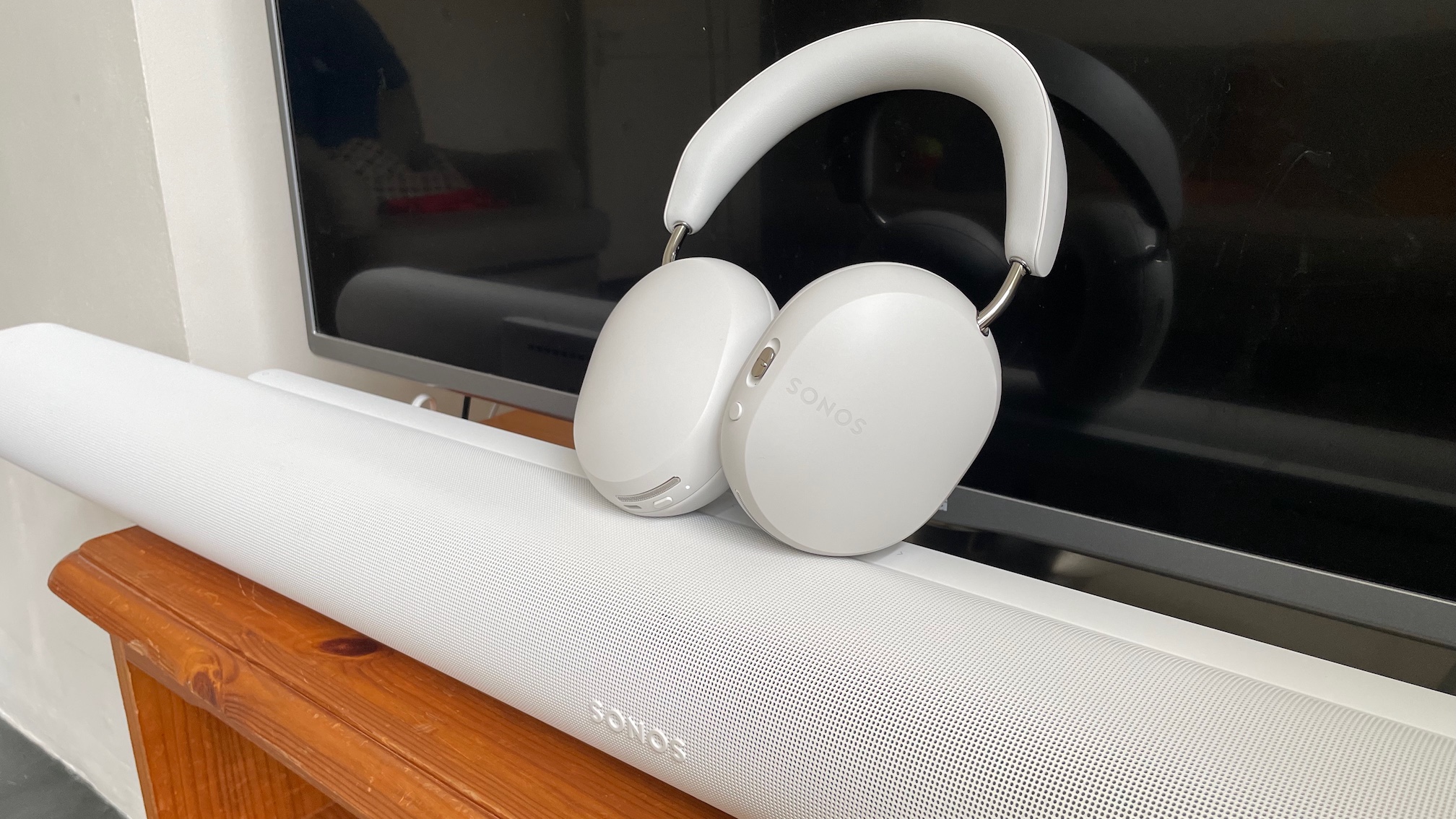 A white pair of Sonos Ace headphones on top of a white Sonos Arc Ultra soundbar in front of a TV.