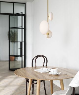 Serene dining room space with low hanging opal and metallic pendant over light wood, round dining table, black dining chair, white painted walls, gray-beige wooden flooring