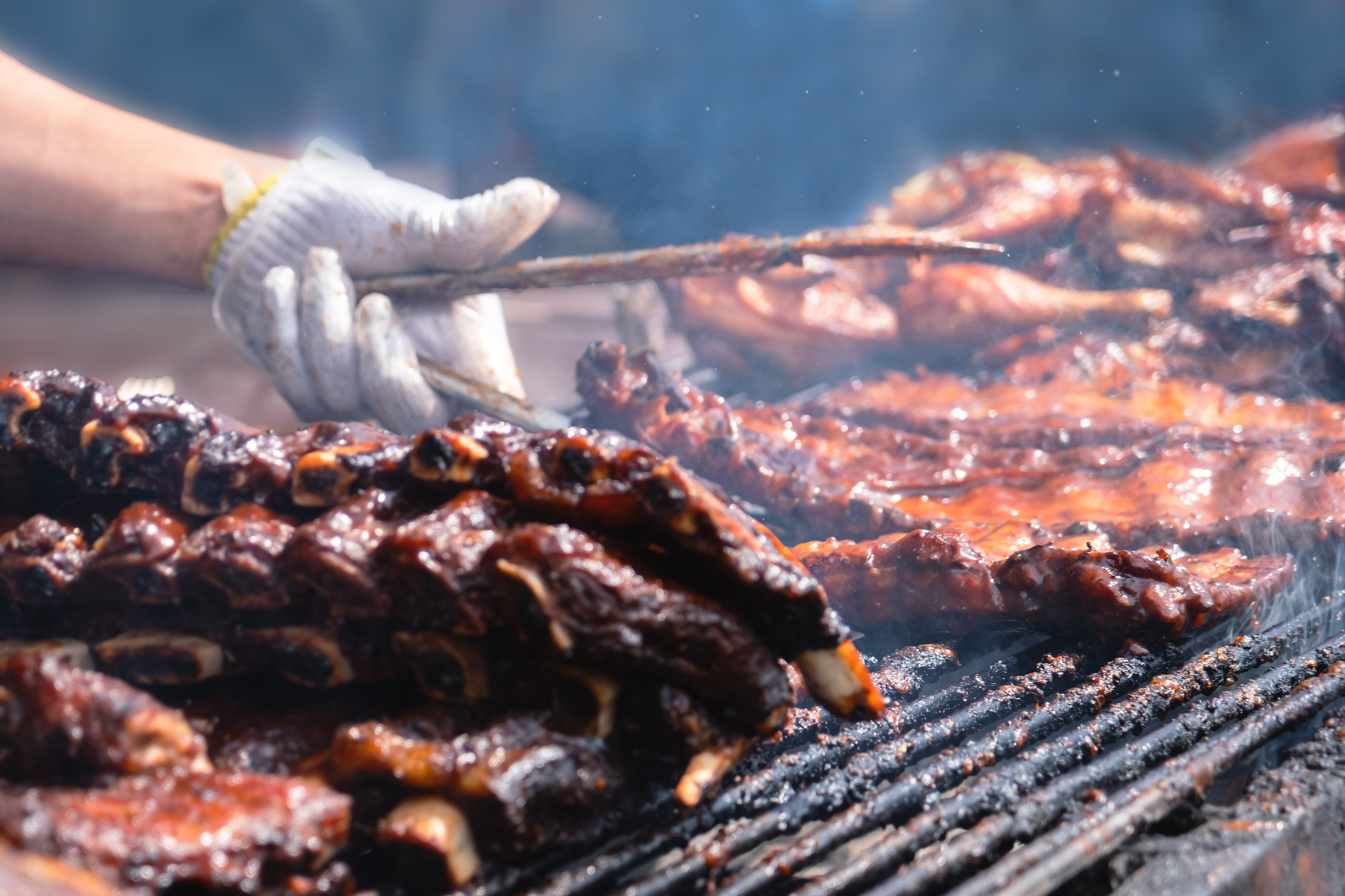 A close-up shot of smoked pork ribs on a BBQ. The arm and hand - in a white glove - are visible, holding a pair of tongs