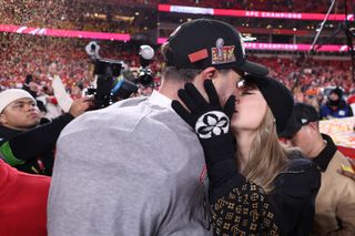 Taylor Swift wearing a black hat, Louis Vuitton patterned sweater and gloves, kissing Travis Kelce on the football field as confetti falls around them at the AFC 2025 Championship game