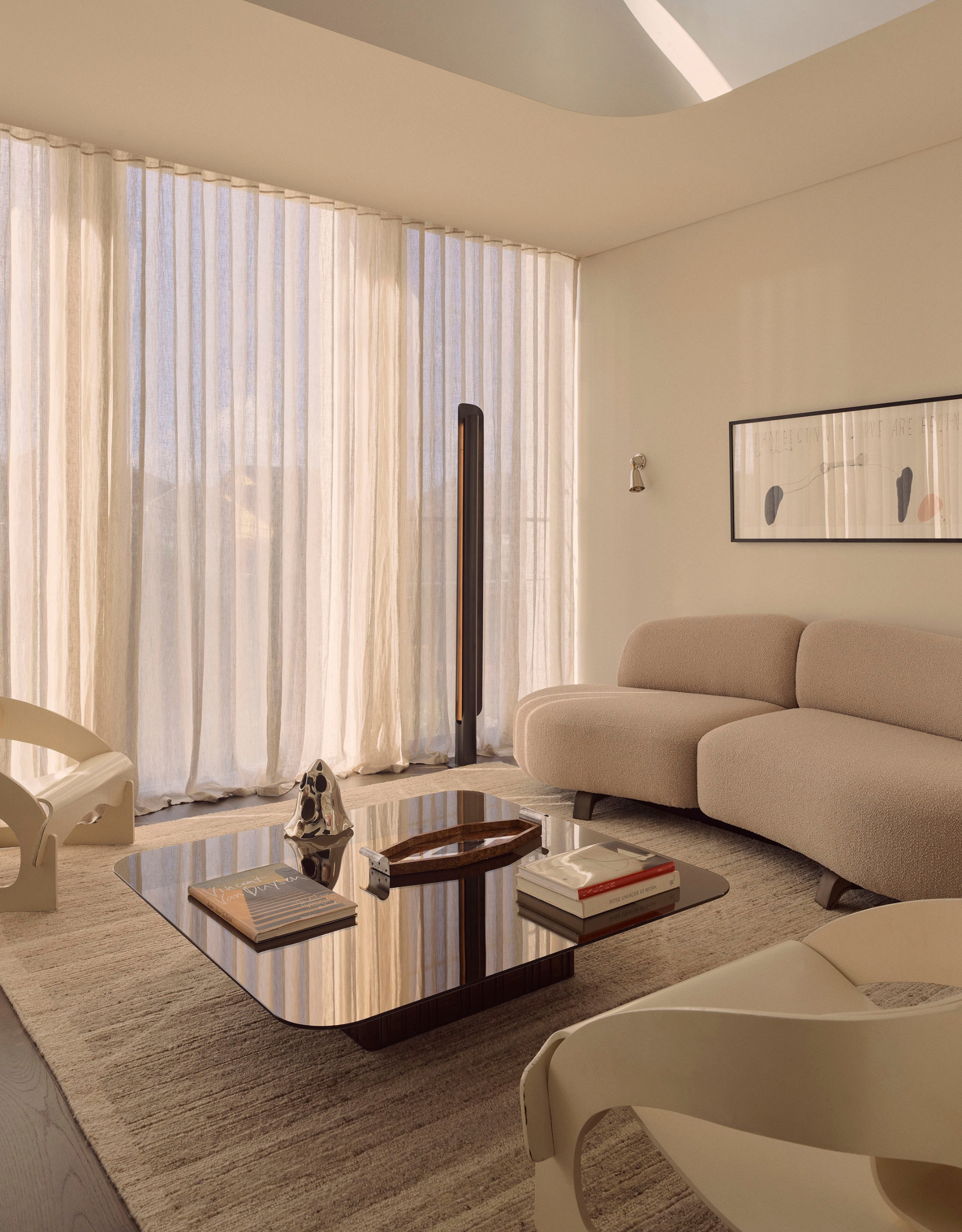 A white living room with a beige couch, a pair of white chairs, a brown acrylic coffee table, and a painting