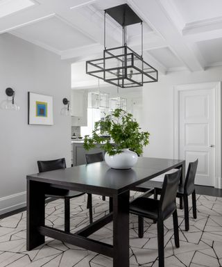 Gray painted dining room with white coffered ceiling paneling, black dining table and four black dining chairs, geometric black metal hanging pendant, plant in white vase on table, wall art and wall lamps mounted on wall, black and cream geometric style rug