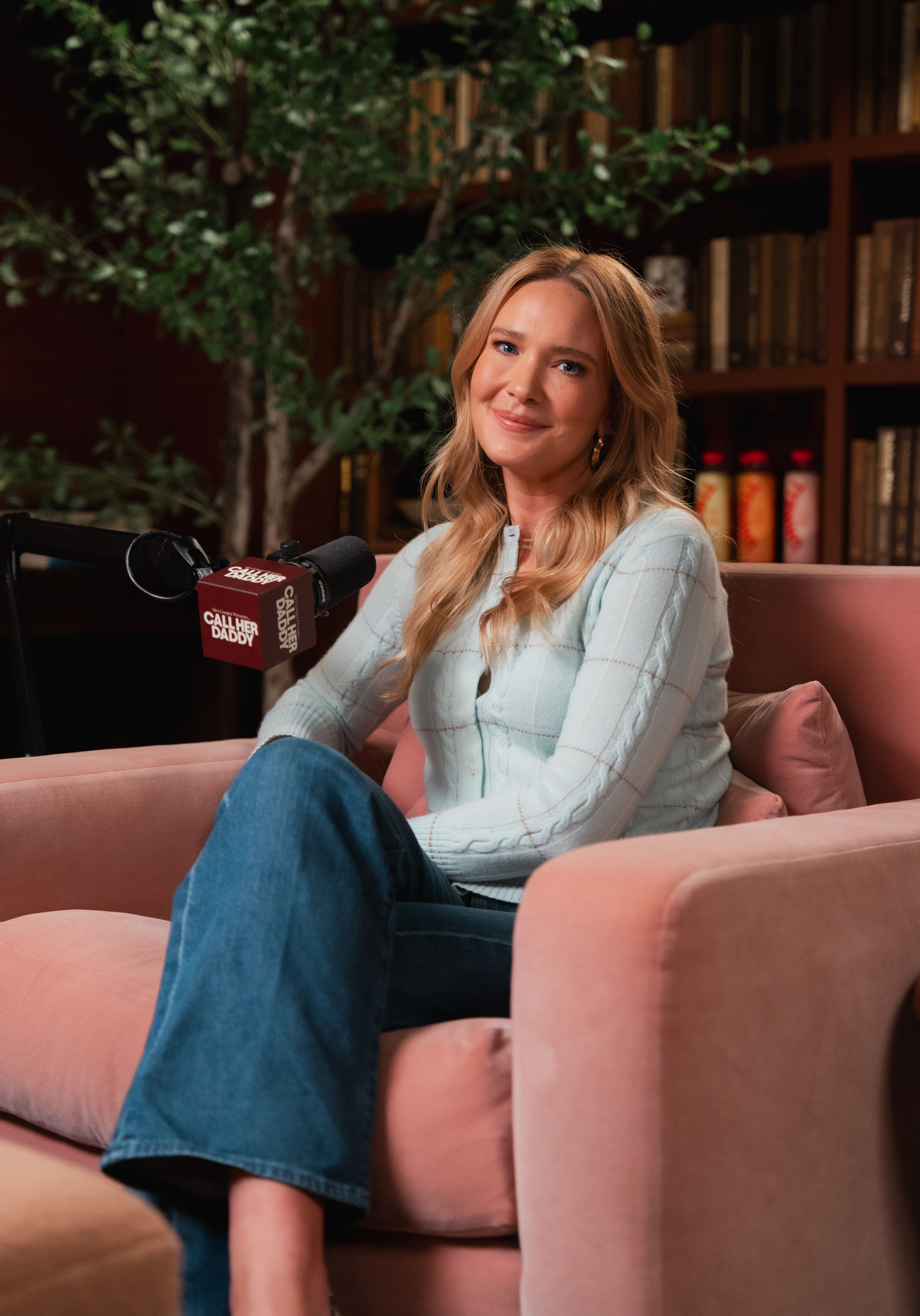 sarah j maas wearing jeans and a blue sweater sitting in a pink chair while appearing on the call her daddy podcast