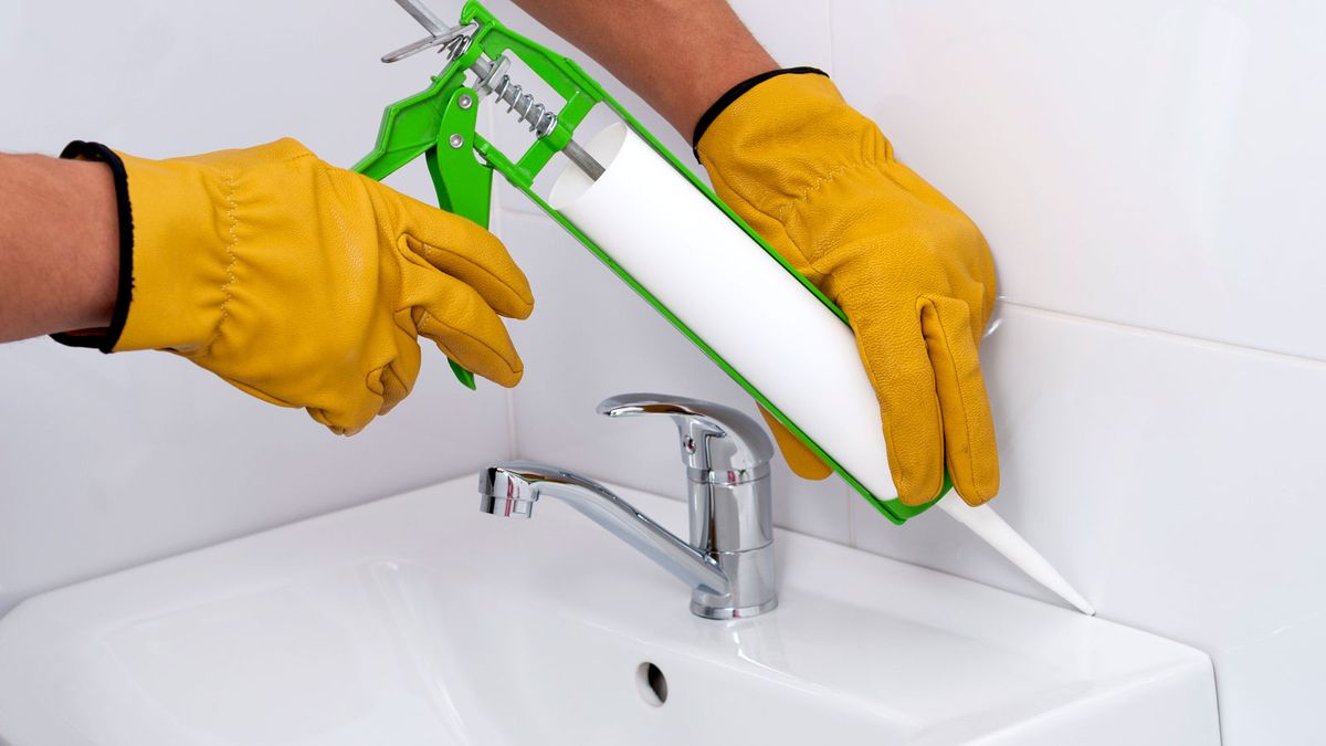 Yellow gloved hands applying bathroom sealant to white tiles and sink