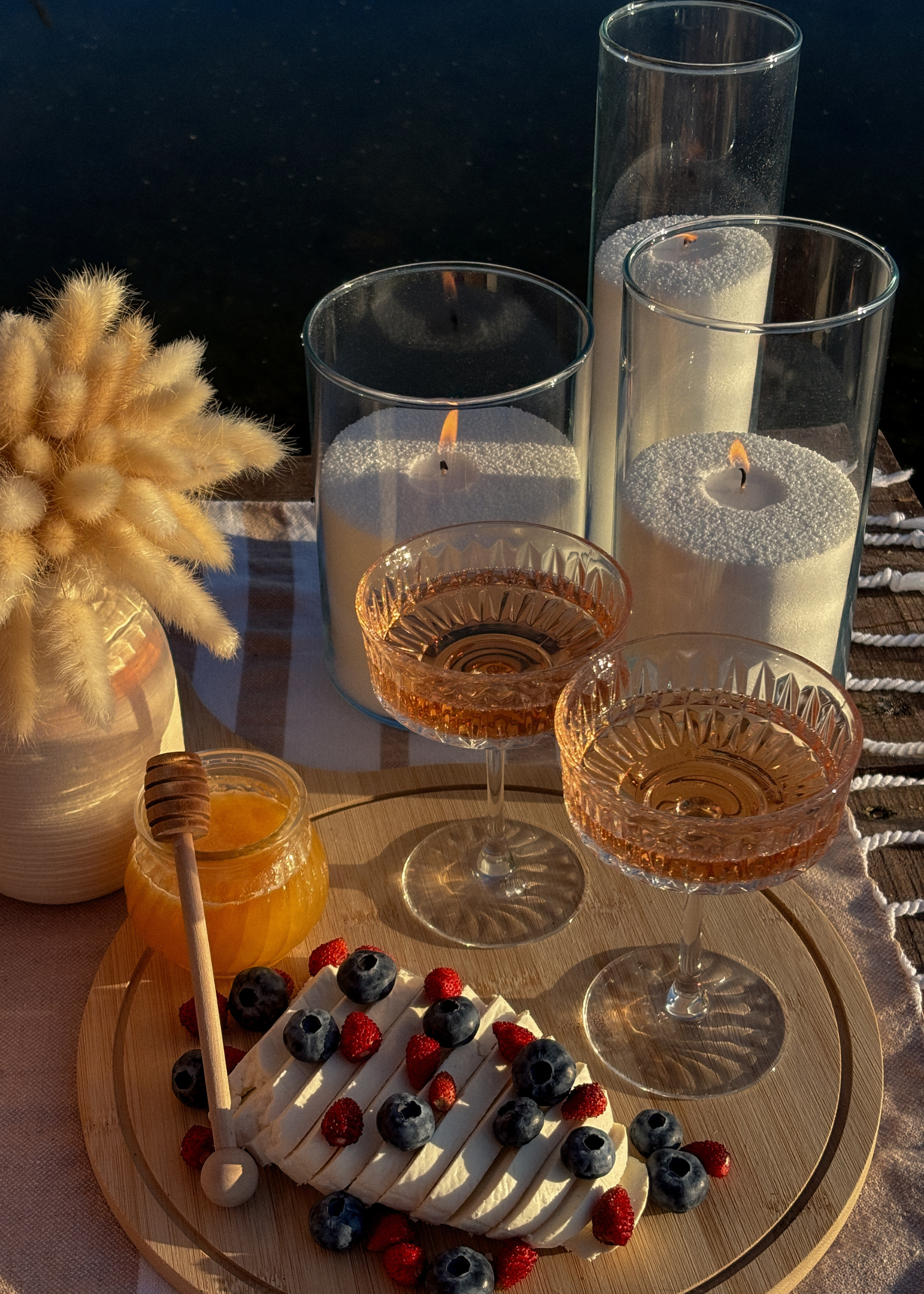 A side table with a vase of fuzzy stems, a wooden board with coupe cocktail glasses, a jar of honey, and berries, alongside three glass cylindrical vases with candle sand