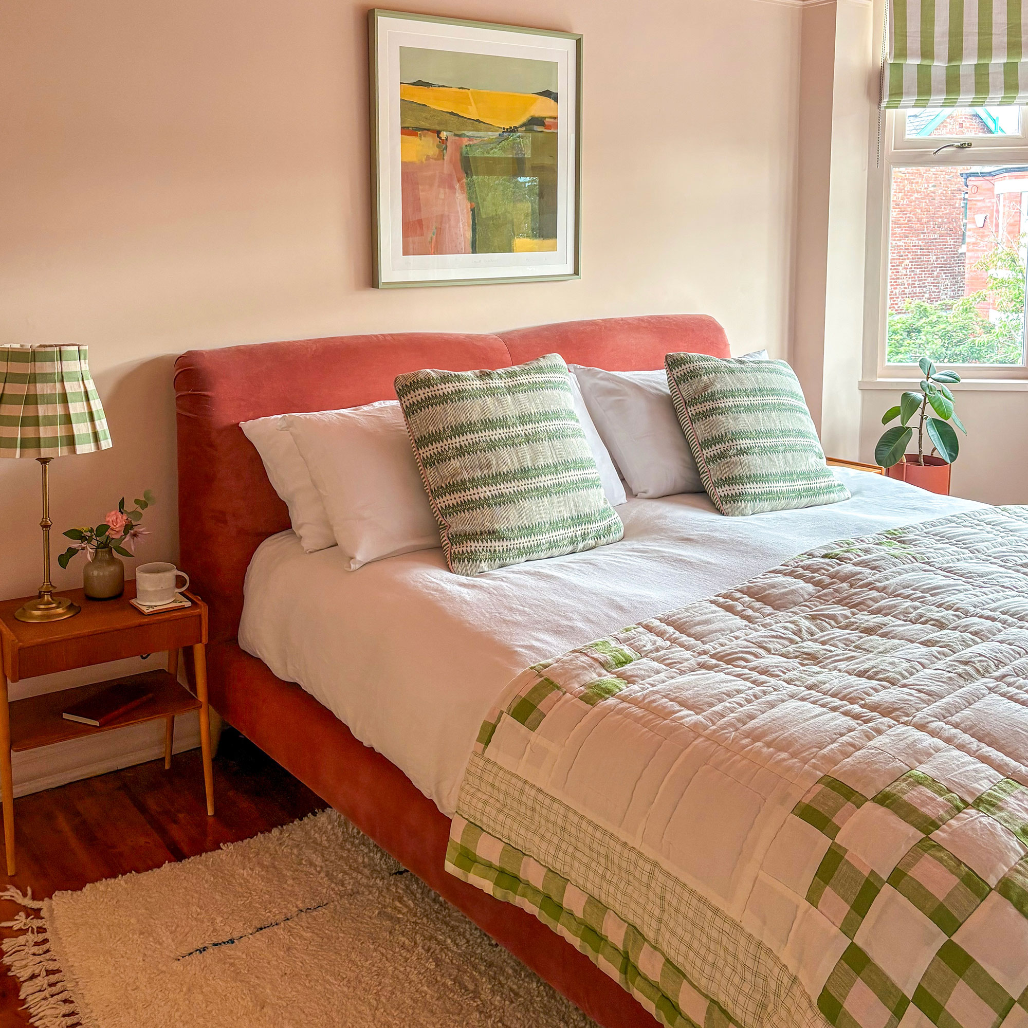 Neutral bedroom with coral coloured upholstered headboard, green and white striped throw cushions, blanket, window blind and lampshade