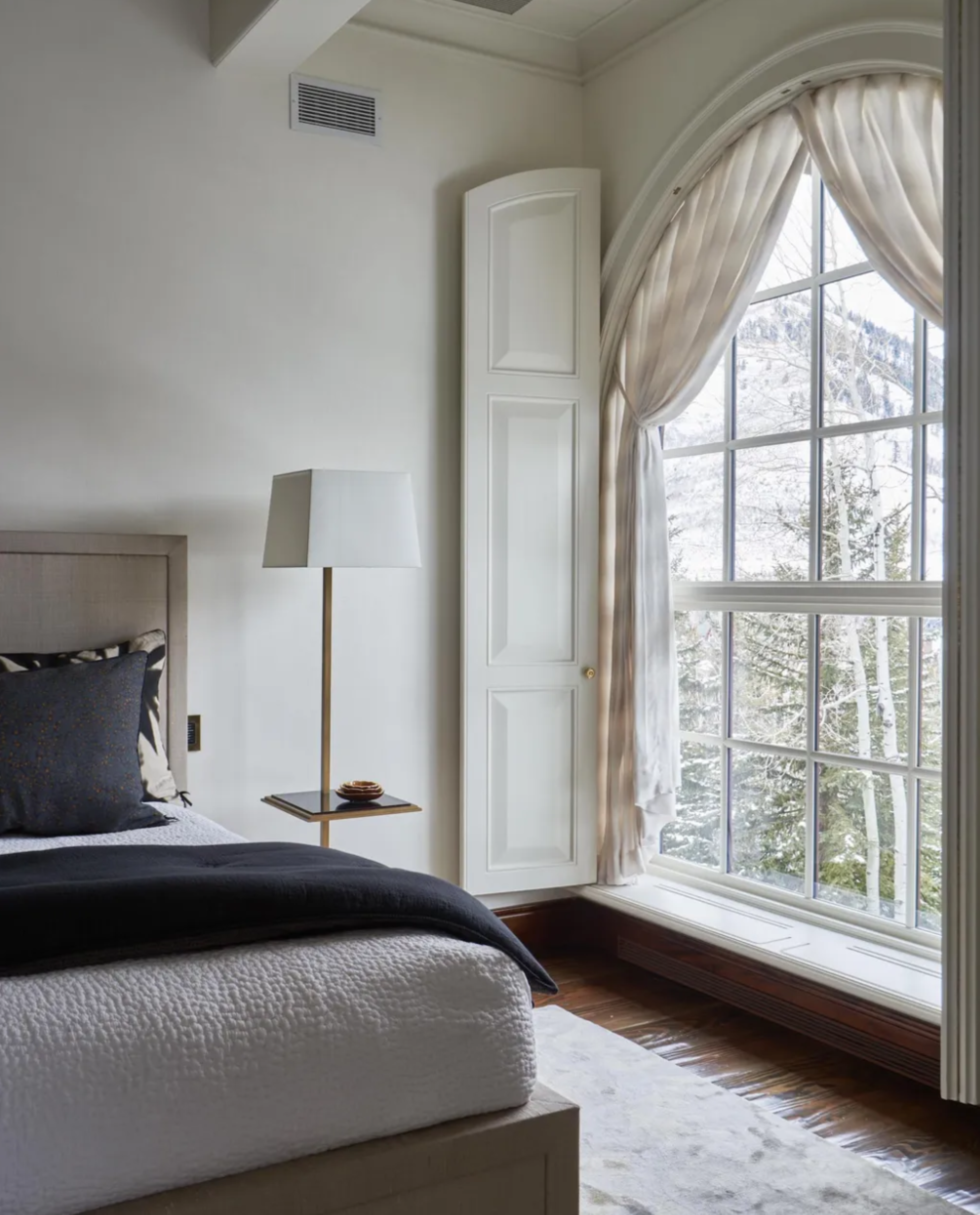 a bedroom with a large window looking outside with off-white curtains pulled back, a hardwood floor covered by a rug, and a tall floor lamp