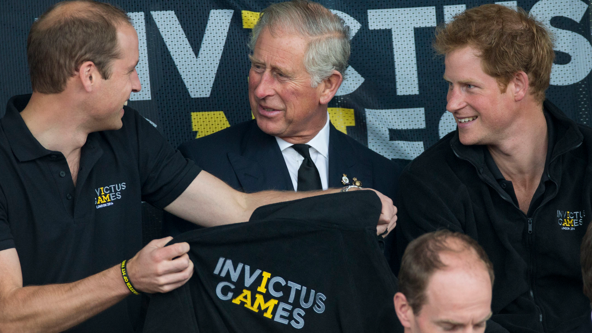 Prince Harry, Prince William and King Charles sitting in front of an Invictus Games banner holding a shirt