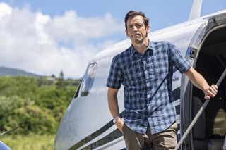 Death In Paradise: Neville (Ralf Little) stands on the steps of a small plane at the airport. He is looking back at the island, his face a mixture of emotions