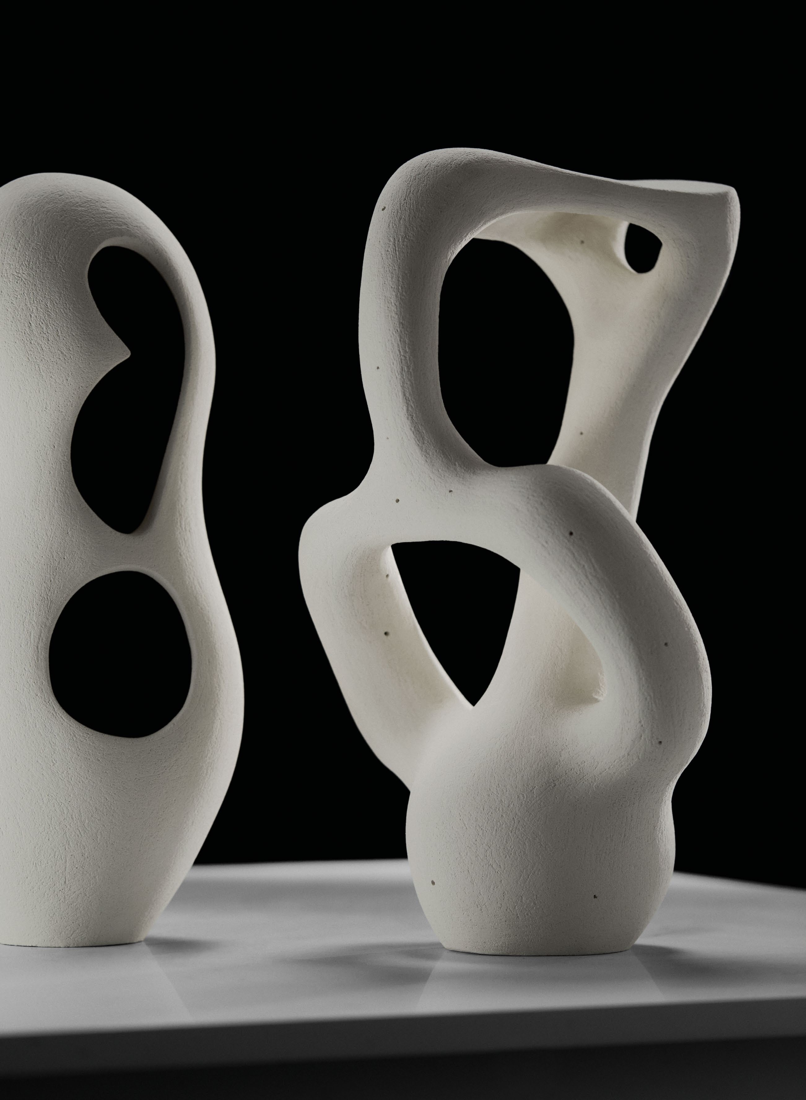 Two abstract white ceramic sculptures by East London-based sculptor Abid Javed, featuring biomorphic forms with smooth, luminescent surfaces.