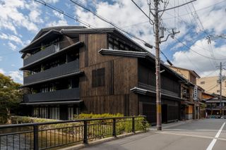 Side exterior view of the black and brown Shinmonzen hotel next to Kyoto's river under a blue cloudy sky