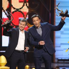 Matt Damon and Ben Affleck on stage together raising their award trophies in celebration.