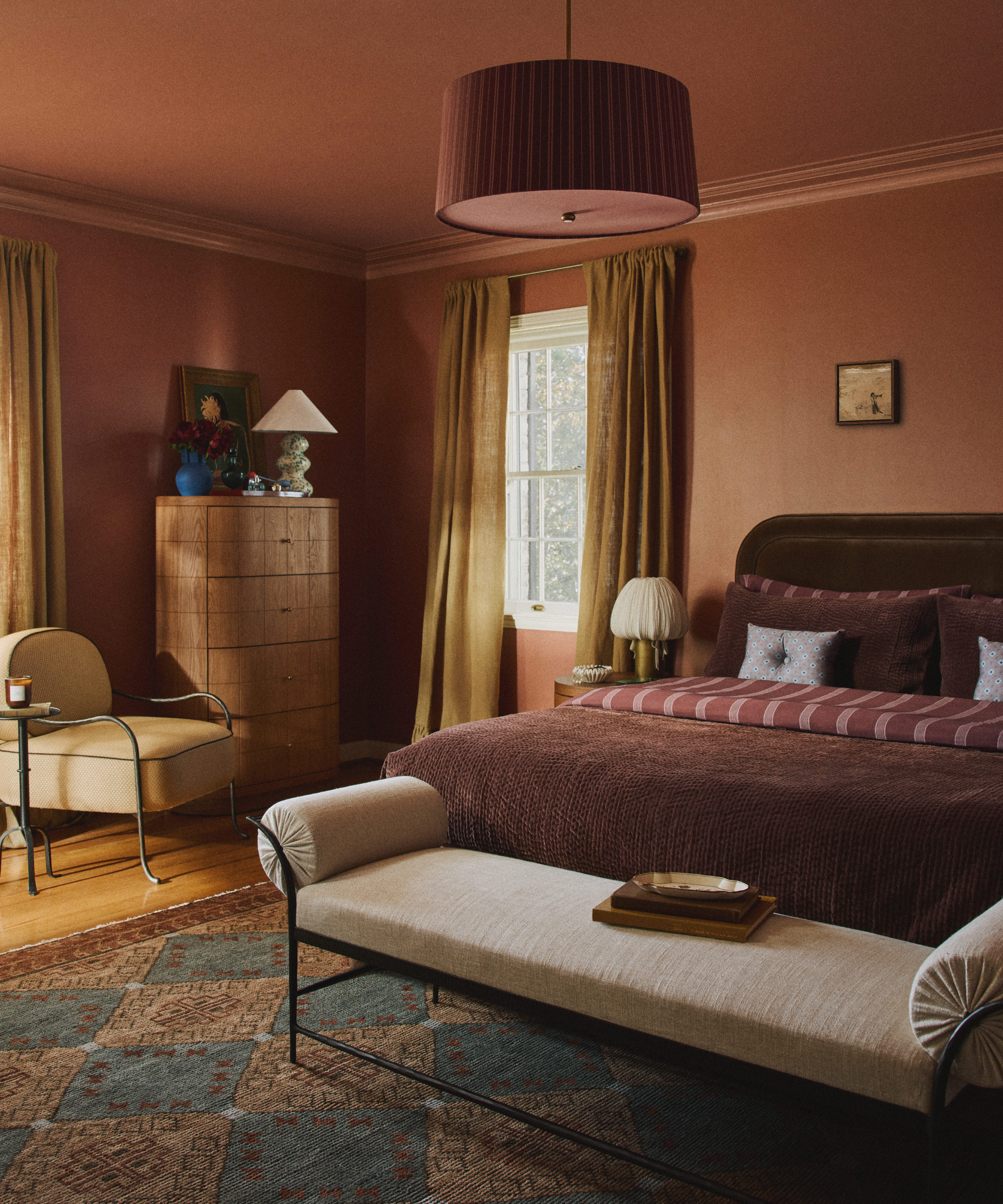 a rust-colored room with a bed with brown bedding, a rust colored pendant, and a tan bench at the foot of the bed