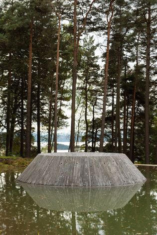 A concrete volcano shaped sculpture in a centre of a man-made pond photographed during the day with tall trees in the background