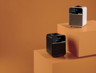 2 bluetooth radio speakers, the one one the left is black and one on the right is white. Both placed on orange boxes with an orange background